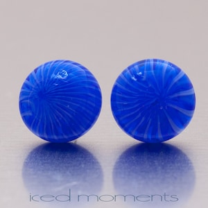 Può includere: Un paio di orecchini in vetro blu con un motivo a spirale bianco. Gli orecchini sono rotondi e hanno una finitura lucida.