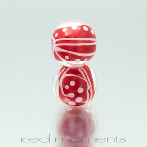 May include: Two red glass beads with white polka dots and stripes. The beads are stacked on top of each other.