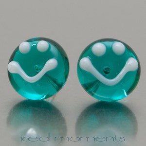 Op de afbeelding: Een paar ronde oorbellen van turquoiseblauw glas met witte smiley-gezichten. De oorbellen hebben een glanzende afwerking.