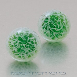 Può includere: Due sfere di vetro verde con vortici bianchi all'interno. Le sfere sono traslucide e hanno una finitura lucida.