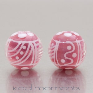 May include: Two pink glass beads with white abstract designs. The beads are round and have a glossy finish.