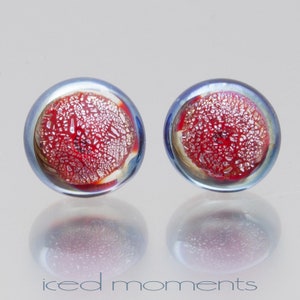 May include: A pair of round glass earrings with a red and white speckled design inside a blue iridescent glass frame.