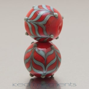 Op de afbeelding: Twee rode glazen kralen met een blauw en zilveren bladmotief. De kralen zijn op elkaar gestapeld.