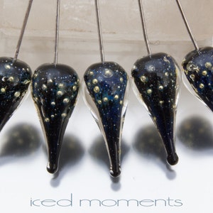 May include: Five teardrop-shaped glass beads with a black and blue marbled pattern and gold flecks. The beads are suspended from thin wires.
