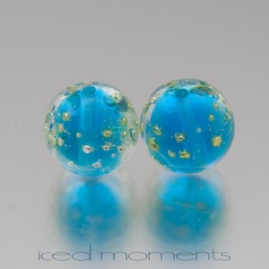 Op de afbeelding: Twee ronde glazen kralen met een lichtblauwe kleur en vlekken van goud en zilver.