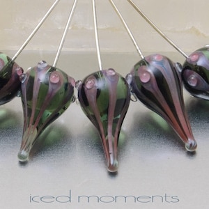 May include: Six glass teardrop-shaped beads with black and green stripes and pink accents are strung on silver pins. The beads are arranged in a semi-circle.