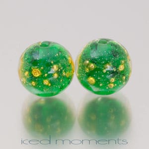 May include: Two green glass beads with gold flecks. The beads are round and have a smooth, shiny surface.