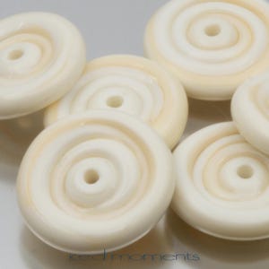 May include: A group of six white and cream colored round beads with a spiral design. The beads have a hole in the center for stringing.