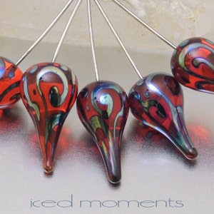 May include: Six red glass teardrop-shaped beads with intricate black and gold swirl designs, each attached to a silver pin.