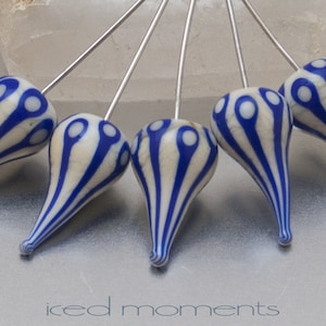 May include: Close-up of several teardrop-shaped beads with a cream-colored base and blue vertical stripes. Each bead has a silver pin and a blue and white pattern at the top. The text "iced moments" is at the bottom.