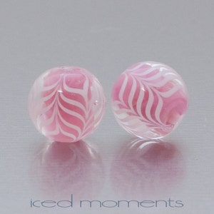 May include: Two clear, round glass beads with a pink and white swirled pattern. The beads have a glossy finish and are set against a light gray background. The text "iced moments" is at the bottom of the image.