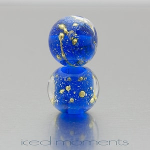 May include: Two blue glass beads with gold flecks. The top bead is round and the bottom bead is slightly flattened. The beads are stacked on top of each other.