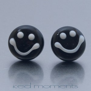 Op de afbeelding: Een paar zwarte oorbel studs met een wit smiley-gezicht.