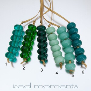 May include: Six strands of glass beads in various shades of green and blue. The beads are arranged in a gradient from dark green to light blue. The beads are strung on a thin brown cord.