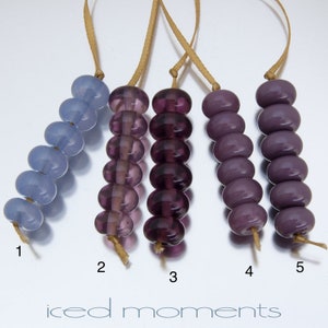 May include: Five strands of glass beads, each with a different color. The beads are strung on a brown cord. The beads are light blue, purple, dark purple, and two shades of dark purple. The beads are labeled 1 through 5.