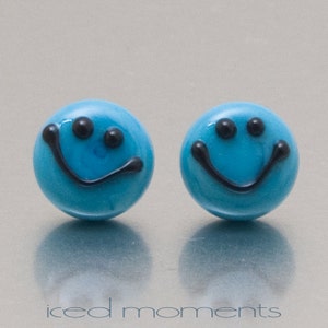 Op de afbeelding: Een paar blauwe glazen oorbellen met zwarte smiley-gezichten. De oorbellen zijn klein en rond.