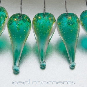 May include: Five teardrop-shaped glass ornaments with a teal blue and gold speckled finish. The ornaments are suspended from a silver wire.