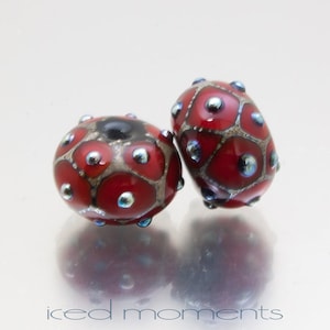 May include: Two red glass beads with a black and silver pattern and blue accents. The beads are round and have a textured surface.