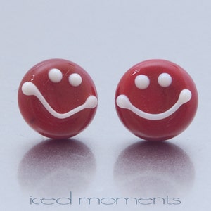 Op de afbeelding: Een paar rode glazen oorbellen met witte smiley-gezichten. De oorbellen zijn rond en hebben een glanzende afwerking.