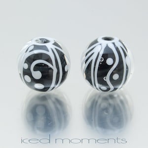 Op de afbeelding: Twee zwarte en witte glazen kralen met een draaiend patroon. De kralen zijn rond en hebben een glanzende afwerking.