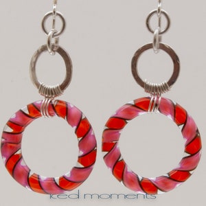 Könnte beinhalten: Silberne Creolen mit pinkfarbenen und roten, gestreiften Glaskreisen. Die Glaskreise sind mit Silberdraht umwickelt.