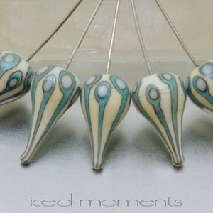 May include: Five teardrop-shaped glass beads with a white and blue swirl pattern are attached to silver pins. The beads are arranged in a fan shape. The text "iced moments" is printed below the beads.
