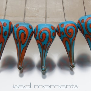 Könnte beinhalten: Fünf türkisblaue gläserne tropfenförmige Ornamente mit orangefarbenen Wirbelmustern, die an silbernen Drähten hängen. Die Ornamente sind in einer Reihe angeordnet.