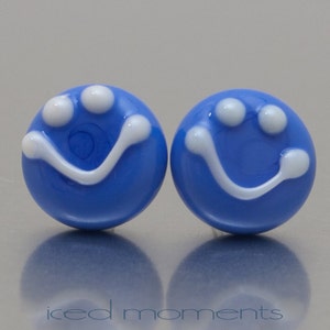 Op de afbeelding: Een paar blauwe glazen oorbellen met witte smiley-gezichten. De oorbellen zijn rond en hebben een glanzende afwerking.