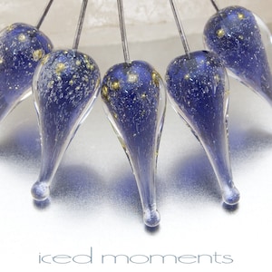 May include: Five teardrop-shaped glass ornaments with a dark blue base and gold flecks. The ornaments are suspended from a wire and hang against a silver background.