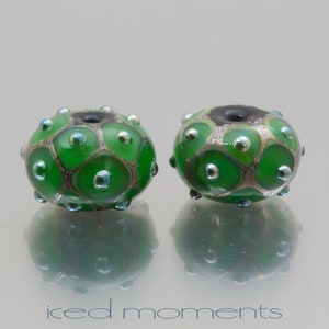 May include: Two green glass beads with silver and black accents. The beads have a raised, textured pattern and are decorated with small, silver dots.