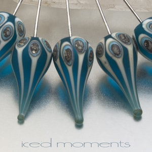 May include: A set of six blue and white glass teardrop-shaped beads with silver accents. The beads are strung on silver wires and are arranged in a fan shape. The beads have a swirling pattern and are reminiscent of a peacock's tail feathers.