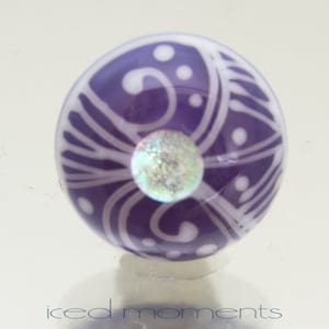 Könnte beinhalten: Lilafarbene Glasperle mit einem weißen Wirbelmuster und einem schillernden Zentrum.