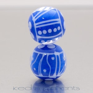 May include: Two blue glass beads with white abstract designs. The beads are stacked on top of each other. The bottom bead is reflected on the surface. The beads have a glossy finish. The text "iced moments" is at the bottom.