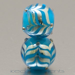 Op de afbeelding: Twee blauwe glazen kralen met een wit en zilver gestreept patroon. De kralen zijn op elkaar gestapeld.