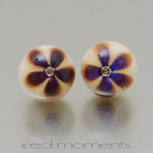 Può includere: Un paio di orecchini rotondi in vetro con sfondo bianco e un motivo a fiore viola. Il centro di ogni fiore ha un piccolo strass trasparente.