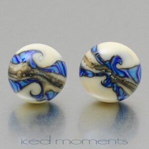 Può includere: Un paio di orecchini rotondi in vetro bianco con vortici blu e marroni. Gli orecchini hanno una finitura lucida.