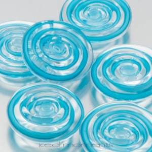 May include: Six clear glass circular ornaments with a swirling blue design. The ornaments are arranged in a semi-circle on a silver surface.