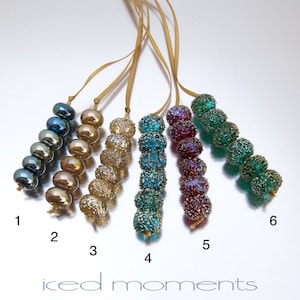 May include: Six strands of iridescent glass beads, each with a different color and pattern. The beads are strung on gold cords and hang from a white background. The beads are numbered 1 through 6.