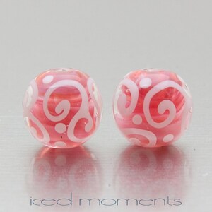 May include: Two pink glass beads with white swirl designs. The beads are round and have a glossy finish.