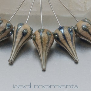 May include: Close-up of five teardrop-shaped beads with silver stems. The beads have a beige, textured surface with dark brown and blue accents. The text "iced moments" is at the bottom of the image.