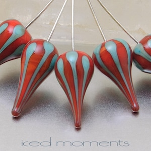 May include: Six teardrop-shaped glass beads with red and teal stripes are attached to silver pins. The beads are arranged in a semi-circle.