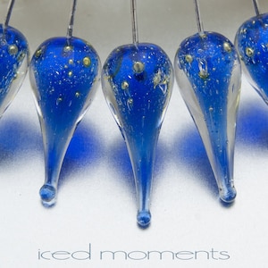 May include: Five teardrop-shaped glass ornaments with a blue and gold speckled design. The ornaments are suspended from a white surface by thin wires.
