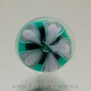 Könnte beinhalten: Ein türkisfarbener Glas-Briefbeschwerer mit einem weißen Blumendesign. Die Blume hat fünf Blütenblätter und eine dunkle Mitte.