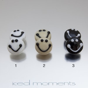 May include: Three pairs of smiley face earrings. The first pair is white with black smiley faces. The second pair is cream with black smiley faces. The third pair is black with white smiley faces.