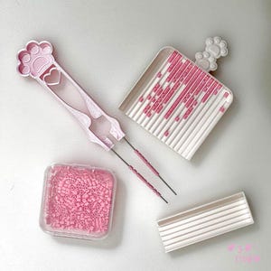 May include: A collection of pink craft tools and supplies, including tweezers with a paw print design, a tray, a container of small beads, and a bead organizer. The tools are designed for crafting and beadwork.