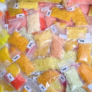 May include: A collection of small, colorful beads in various shades of yellow, orange, pink, and white, packaged in clear plastic bags. Each bag is labeled with a white tag, featuring a letter and number combination, such as "A26" or "A9".