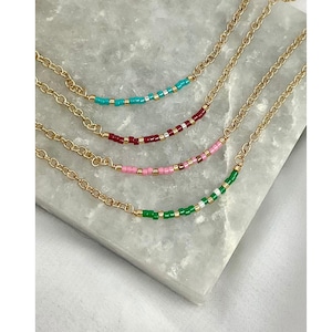 May include: Four gold chain necklaces with different colored beaded sections. The necklaces are layered on top of each other, with the green beaded section at the bottom, followed by pink, red, and turquoise.