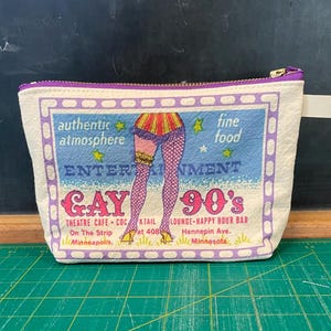May include: A white canvas zippered pouch with a purple trim. The pouch features a colorful illustration of a woman's legs in fishnet stockings and a red and yellow skirt. The text "Gay 90's" is printed in large, bold letters. The text "Entertainment" is printed above the text "Gay 90's". The text "Authentic atmosphere" is printed in the top left corner of the pouch. The text "Fine food" is printed in the top right corner of the pouch. The text "Theatre Cafe Cocktail Lounge Happy Hour Bar" is printed below the text "Gay 90's". The text "On The Strip Minneapolis" is printed below the text "Theatre Cafe Cocktail Lounge Happy Hour Bar". The text "at 408 Hennepin Ave. Minnesota" is printed below the text "On The Strip Minneapolis".