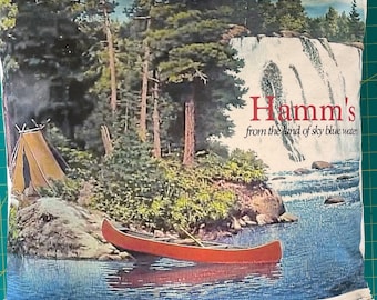 May include: A white pillow with a zipper closure features a vintage-style image of a waterfall, a canoe, and a tent in a forest setting. The text "Hamm's from the land of sky blue water" is printed in red on the pillow.
