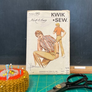 May include: Vintage Kwik Sew pattern 910, featuring a retro illustration of a man in a shirt and pants. The pattern is accompanied by a pin cushion with pins and a pair of black scissors on a green cutting mat.